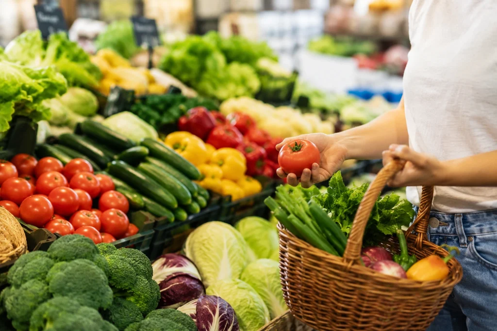 Person selecting fresh vegetables and tomatoes at a grocery market with a basket of produce